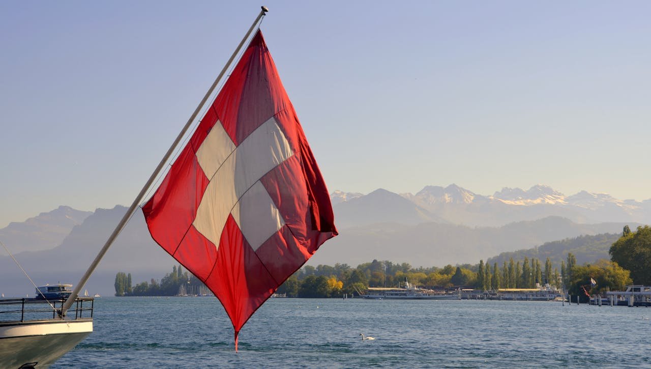 Swiss flag on a flagpole near a serene lake with breathtaking mountain backdrop in Switzerland.